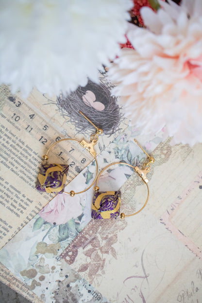 A pair of origami Sonobe earrings displayed on a table with a floral pattern, alongside a grey fabric and an open book with Japanese text.