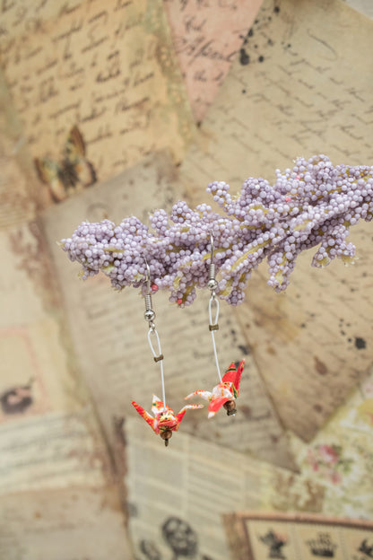 A pair of handmade origami crane dangle earrings made from red and white chiyogami paper, hanging from a purple flower stem and displayed against a background with paper and text.
