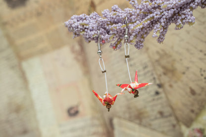 A closeup of a pair of handmade origami crane earrings made from red and white chiyogami paper, hanging from a flower against a background with handwritten papers.