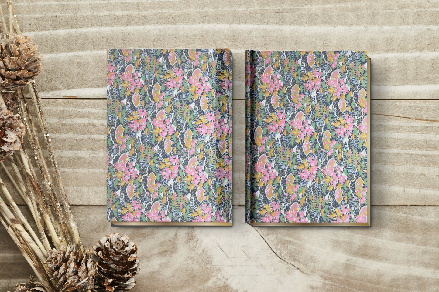 Two floral-patterned notebooks made with chiyogami paper in a bookbinding project on a wooden surface with pine cones.