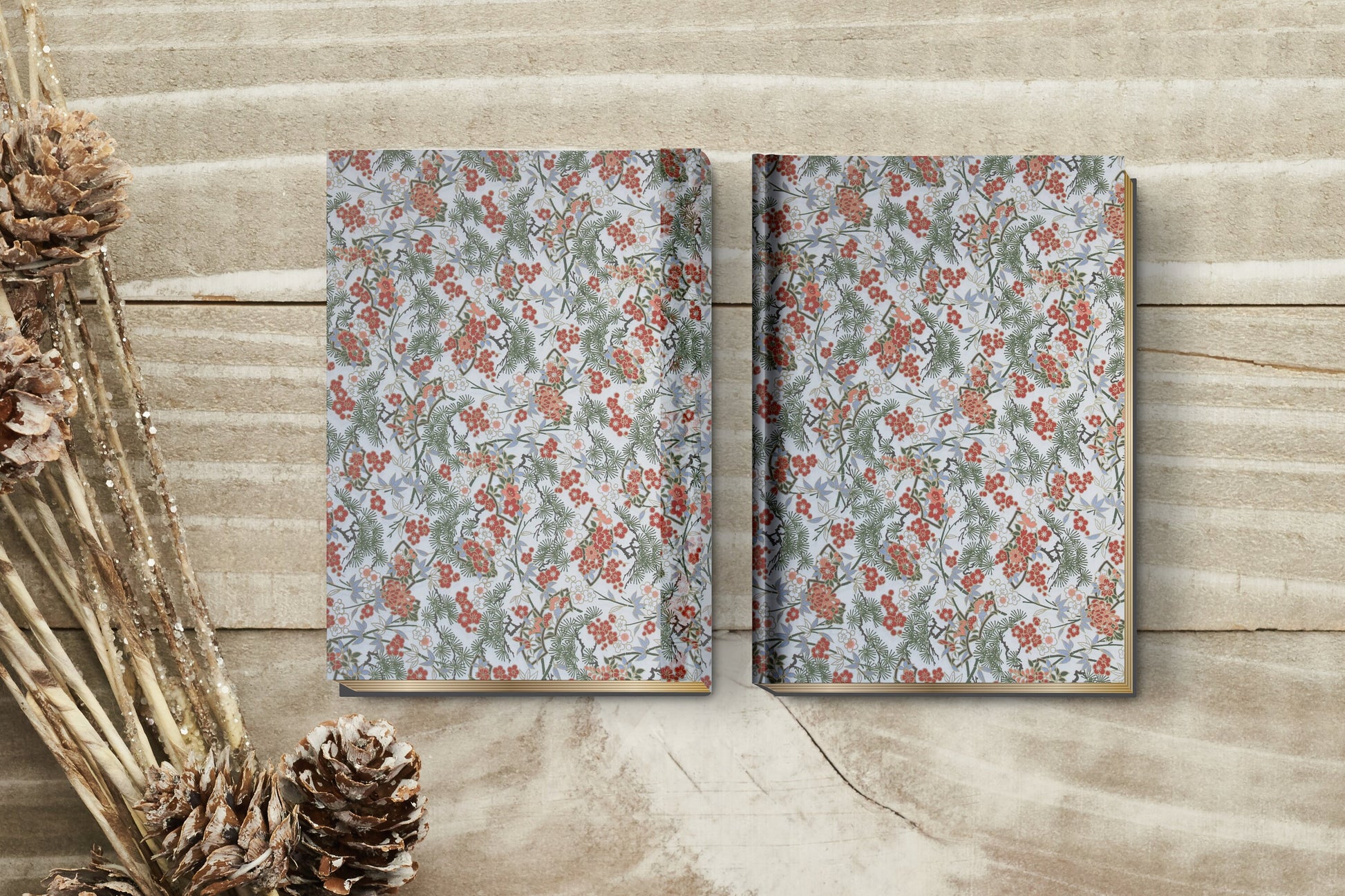 Two floral-patterned notebooks made with chiyogami paper in a bookbinding project on a wooden surface with pine cones and branches.