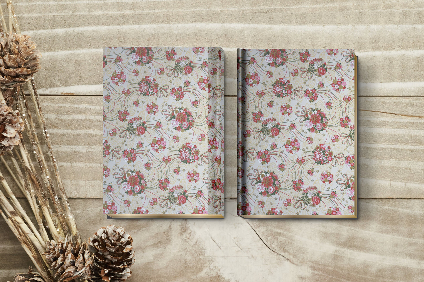Two floral-patterned notebooks on a wooden surface with pine cones and branches.