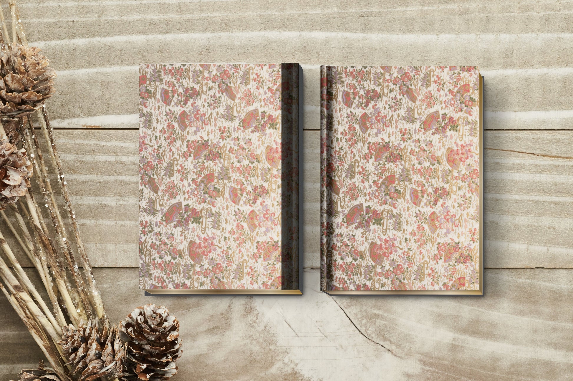 Two notebooks with floral and leaf patterned with red, green, and brown colors wrapped with chiyogami paper on a wooden surface with pine cones.
